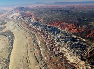 Oblique aerial photograph of a long line of multicolored rock beds dipping into the ground. The beds are fractured and erode in a way that makes the parts sticking out look like triangles.