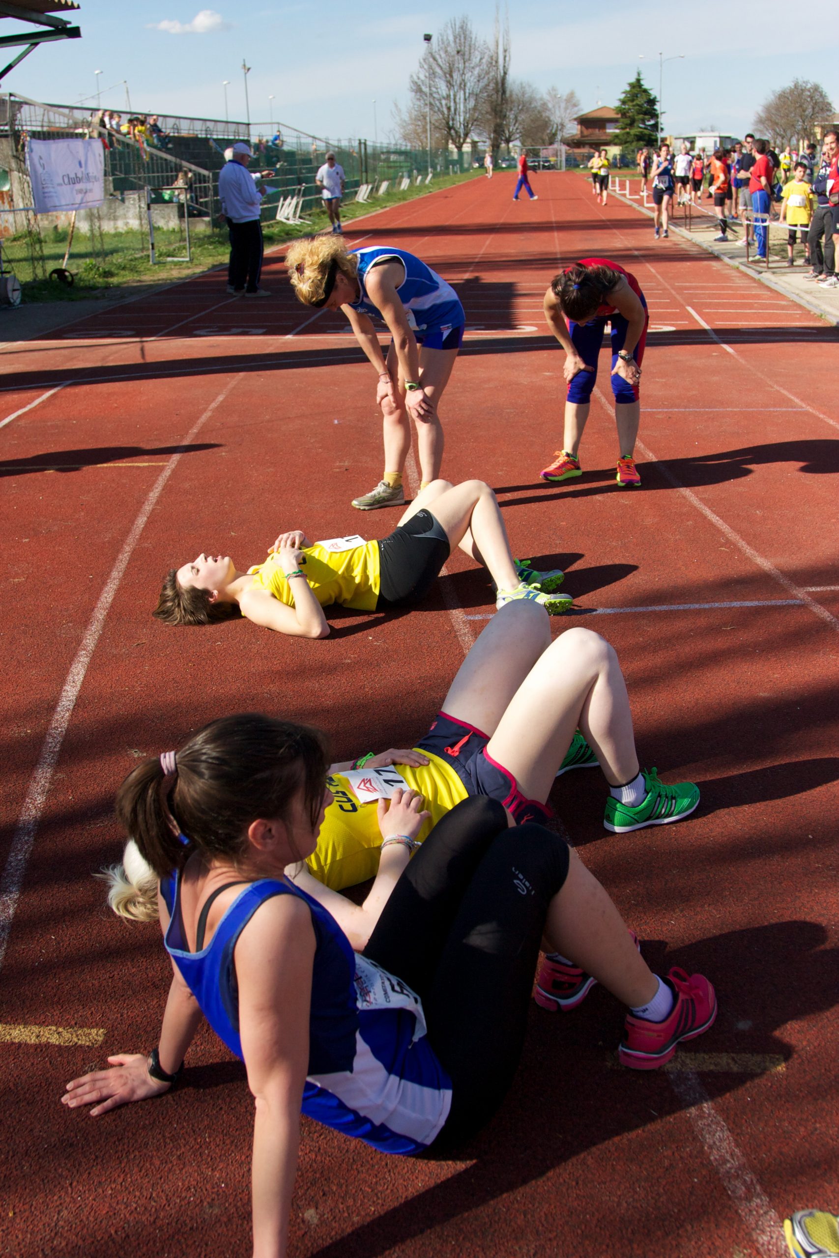 Image shows female track and field runners resting after a race. Three women are resting on the ground and two are leaning over with their hands on their knees, catching their breathe.