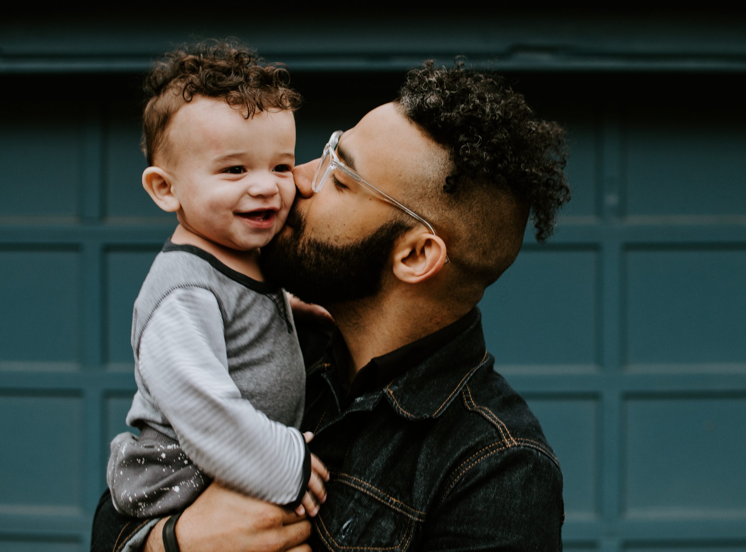 Image shows a dark, curly-haired man in his 20s or 30s holding and kissing a toddler with similar physical features and curly, dark hair, while the toddler smiles. 