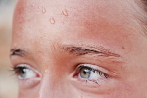 Image shows a close-up view of the upper portion of a person's face. The person's skin shows redness due to heat and beads of sweat on their brow.