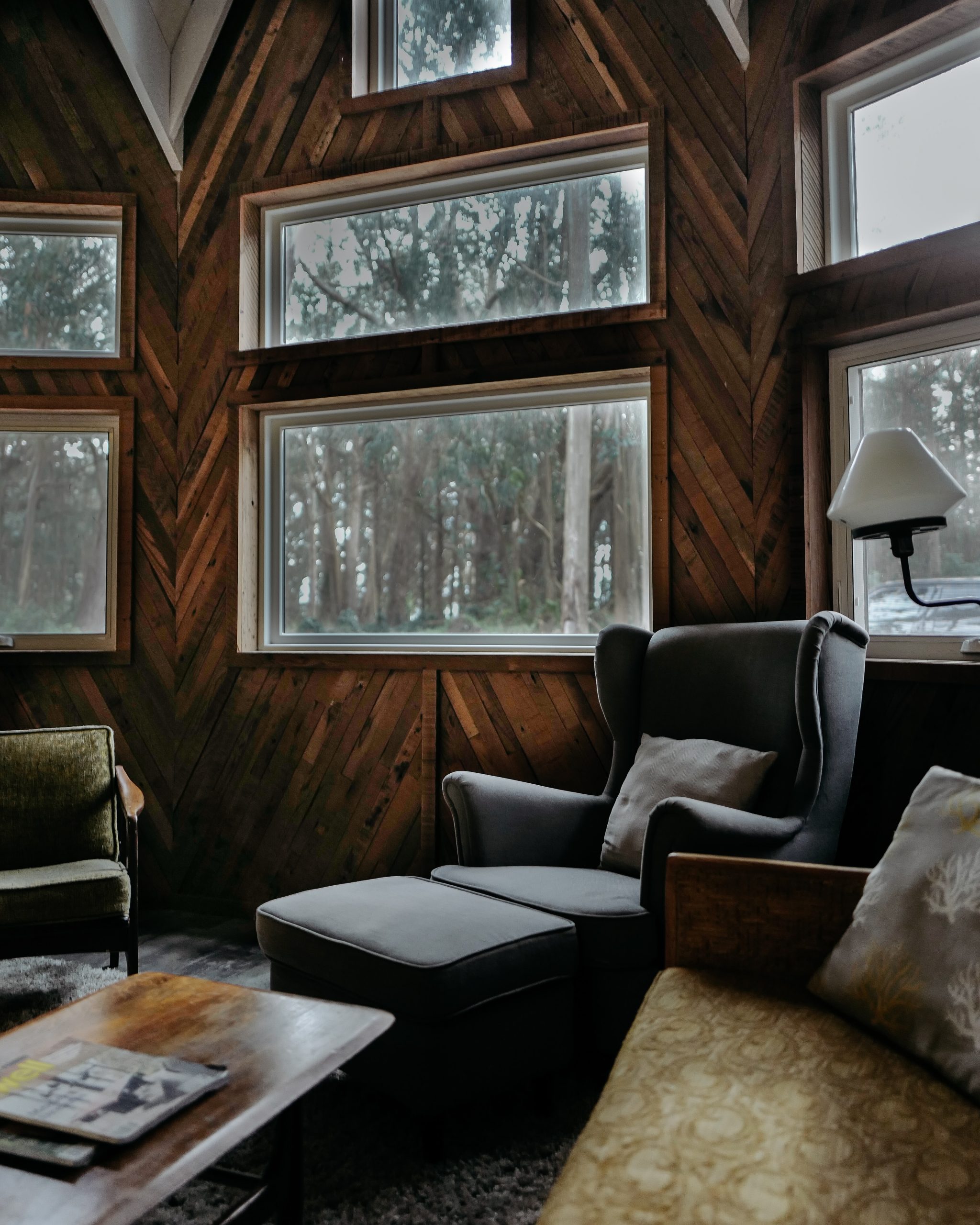 Image shows a photo of a living room with large windows. There is a leather armchair, coffee table, lamp and books. The walls have wood panelling.