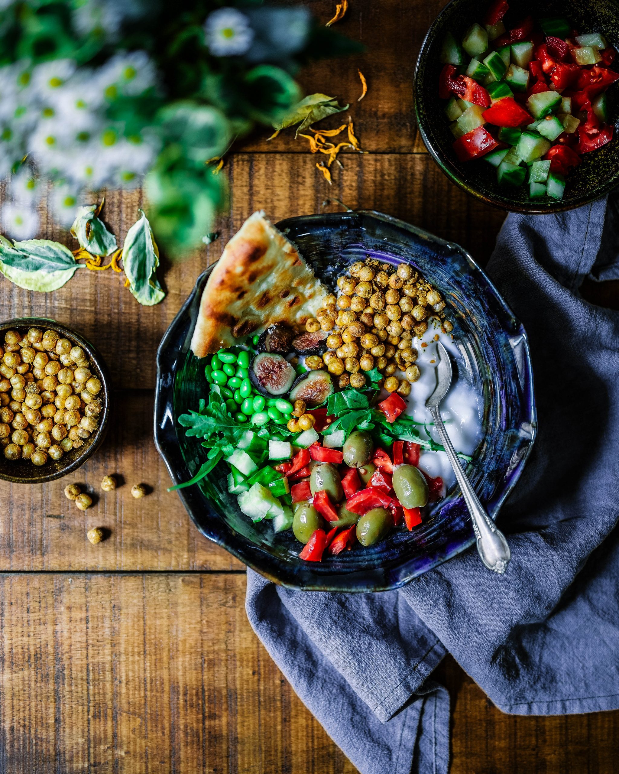 Image shows a blue plate holding yogurt, soy beans, olives, pimentos, chickpeas, flatbread and various other diced vegetables.