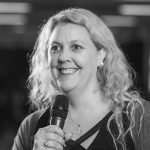 Black and white photo of Betsy. She is a blonde, middle-aged white woman and she is holding a microphone and smiling