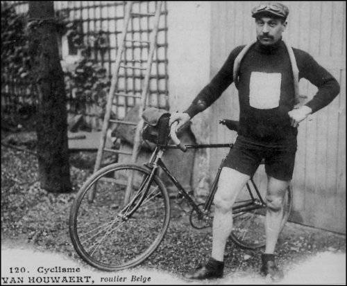 Cyrille Van Hauwaert standing next to an early 20th-century bicycle. He is dressed in black shorts, a long-sleeved shirt, with a bag on his back.