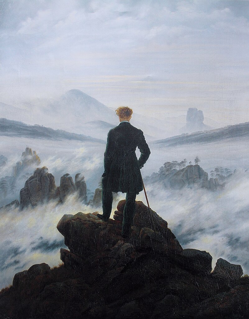 Figure of a man wearing dark clothing and light hair standing with is back to the viewer on a ledge of rocks overlooking a valley of fog.