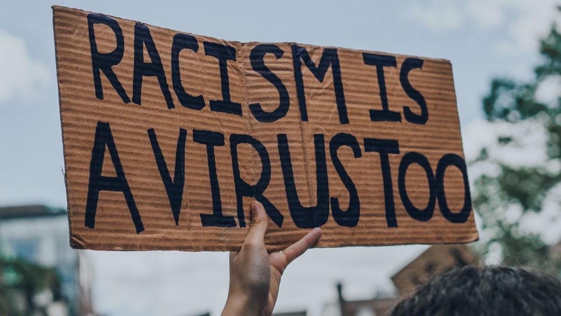 A placard that says "Racism is a virus too", held by a black hand with a tree at the back.