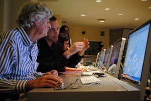A younger man and older man sitting at a computer with the younger man explaining how a computer program works.