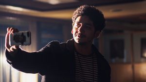 A man wearing a striped shirt and cardigan with half of his face hidden in shadow taking a selfie.