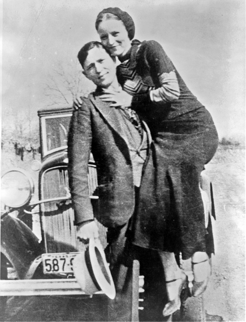 Bonnie and Clyde standing in front of a car. Clyde holding Bonnie in a seated position, holding his hat in the other.