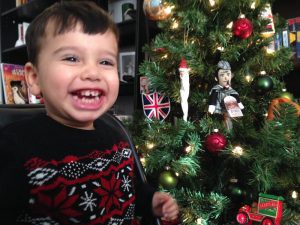 A young darked haired by wearing a Christmas sweater smiles next to a decorated Christmas tree.