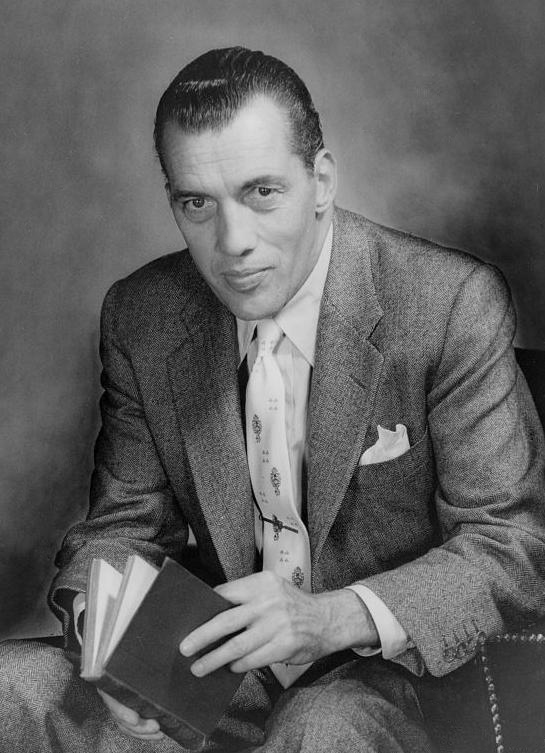 Portrait of Ed Sullivan sitting in a chair holding a book, looking at the camera.
