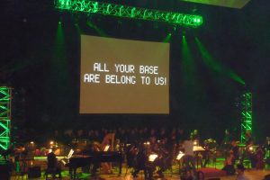 "All Your Base Are Belong to Us" displayed on a large screen during an orchestral performance.