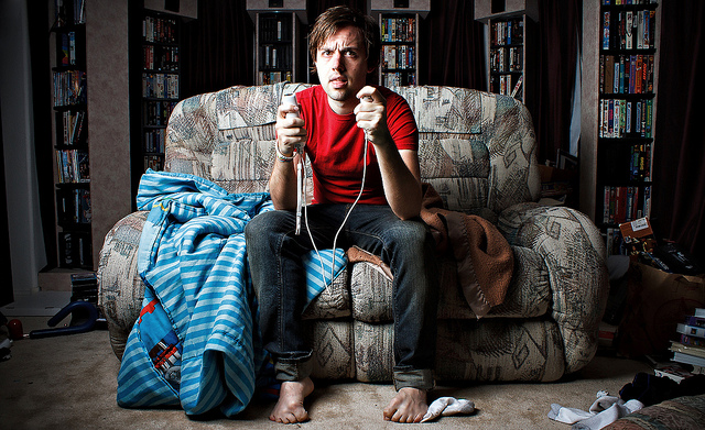 Man sitting on messy couch holding video game controllers in each hand.