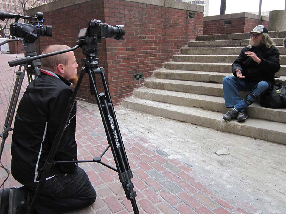 Filmmaker sitting behind a camera as he interviews a man sitting on outdoor steps.