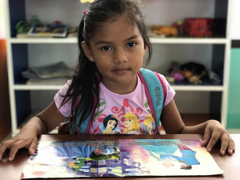 Preschool girl sitting at a table wearing a backpack reading a book on the table in front of her.