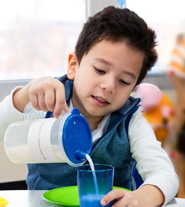 Four year old boy pouring milk from a pitcher into a cup.