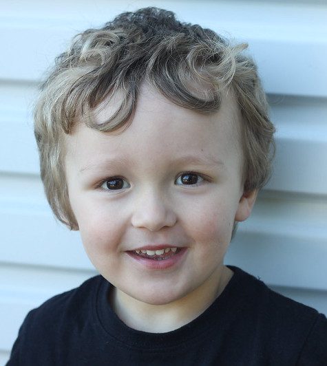 Blond two year old standing in front of a wall smiling.