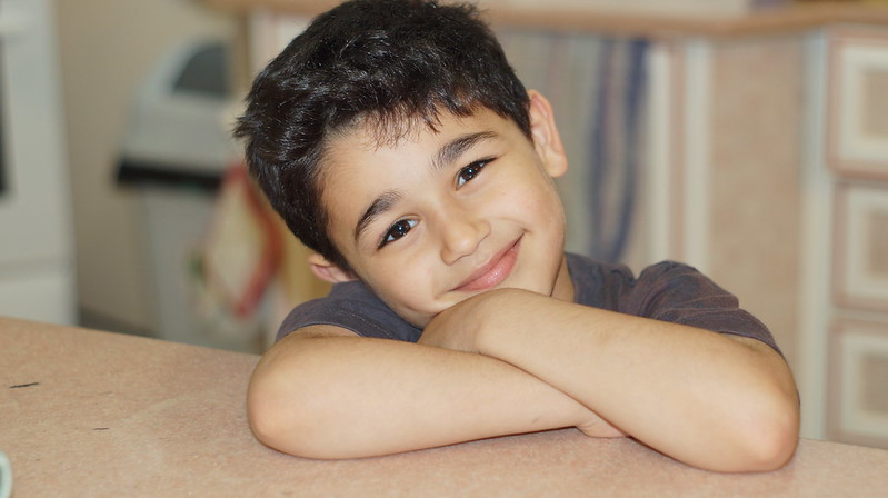 Boy with head leaning on crossed arms on a table smiling.