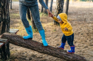 Woman in blue jeans with blue rain boots holding the hand of a little boy in a yellow hoodie walking up a wood log.
