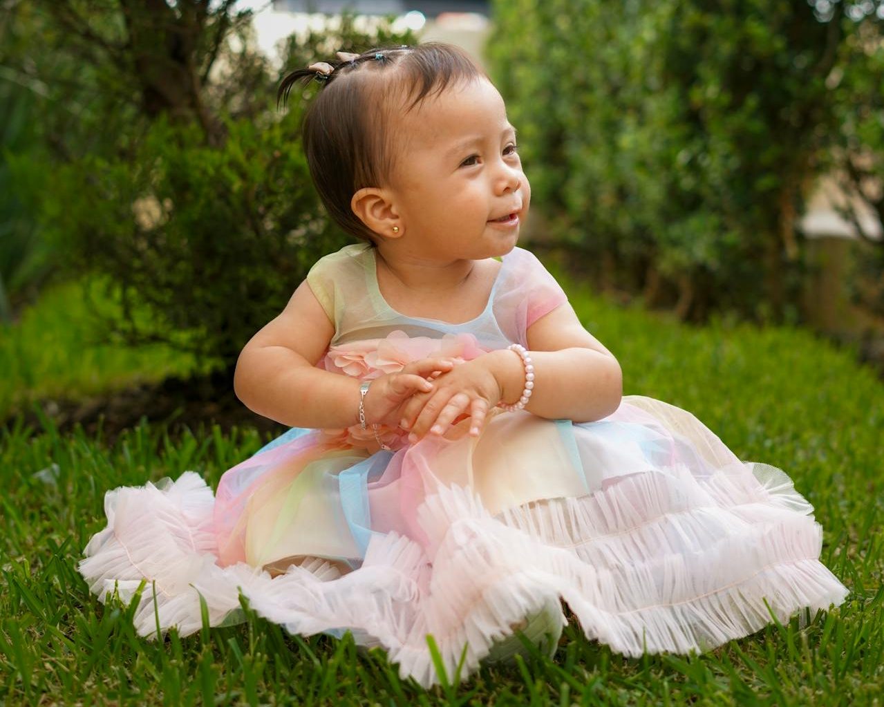Toddler girl sitting on grass in pastel rainbow dress.