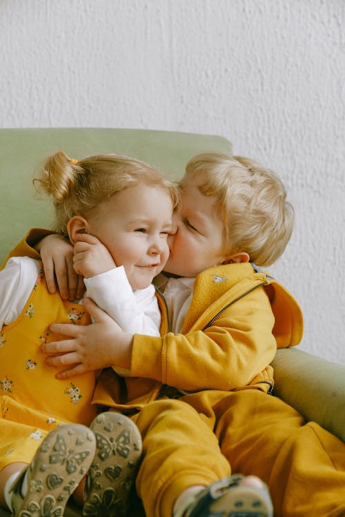 Toddler brother and sister in yellow clothes with blonde hair sitting in a green armchair hugging. The brother is kissing the sister on the cheek.