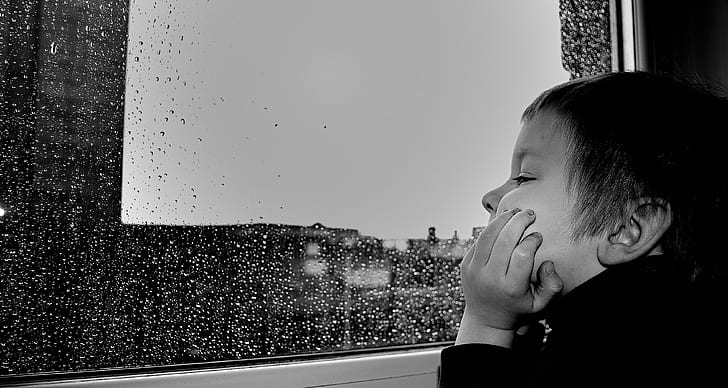 Grayscale photo of young boy watching rain through the window.