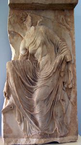 A woman whose body is revealed through her robes as she adjusts her sandal.