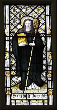 A stained-glass window illustrating Hildegard of Bingen standing with a golden book and holding a staff. Her sainthood is indicated by a halo.