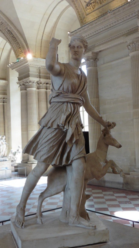 A stone sculpture of a woman whose dress falls above her knees. She holds the horn of a rearing stag in her left hand. Her right hand is raised. Her hair is pulled back and she wears sandals.