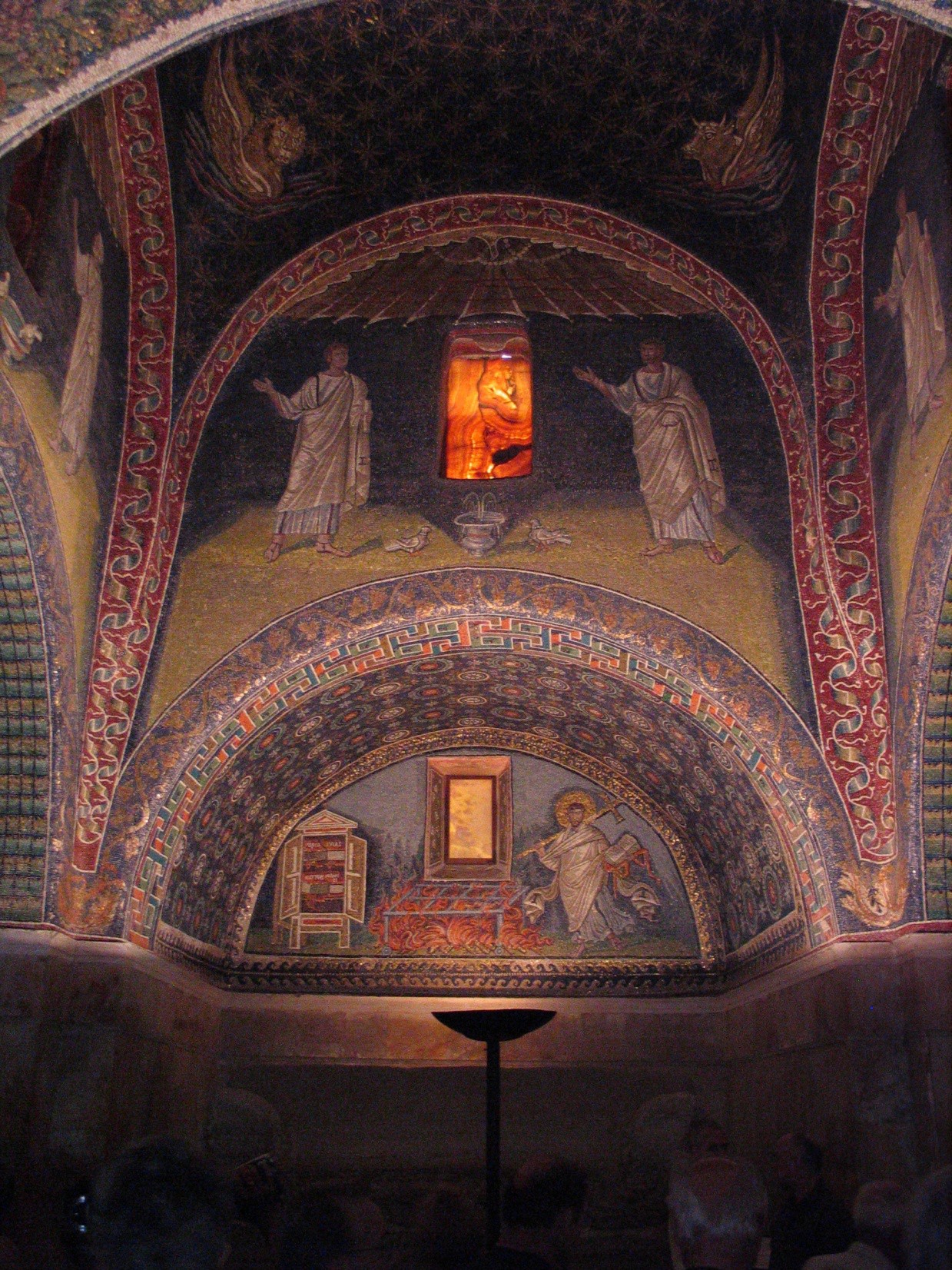 A mosaic depicting a man in a robe moving toward a fire with a cross over his shoulder. He carries a bible in his left hand. Above this is a mosaic depicting two men and two doves around a small fountain.