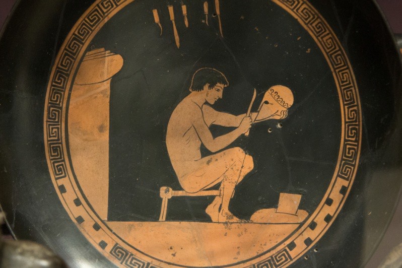 An image on red figure pottery. It is an idealized nude craftsman working on a helmet. Geometric designs run around the rim of the plate.