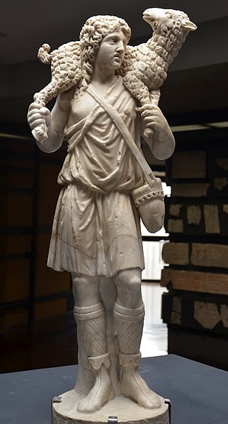 A carved shepherd carrying a sheep over his shoulders. He has curly hair a short tunic and boots.
