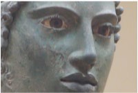 A close-up of the face of a bronze statue. The face is ideal but natural with inset eyes and eyelashes. Curly sideburns are apparent on his otherwise smooth face.