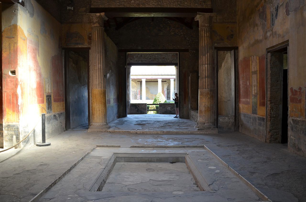 A large room held up by pillars with a shallow pool in the center and an open doorway at the end, past the pillars.