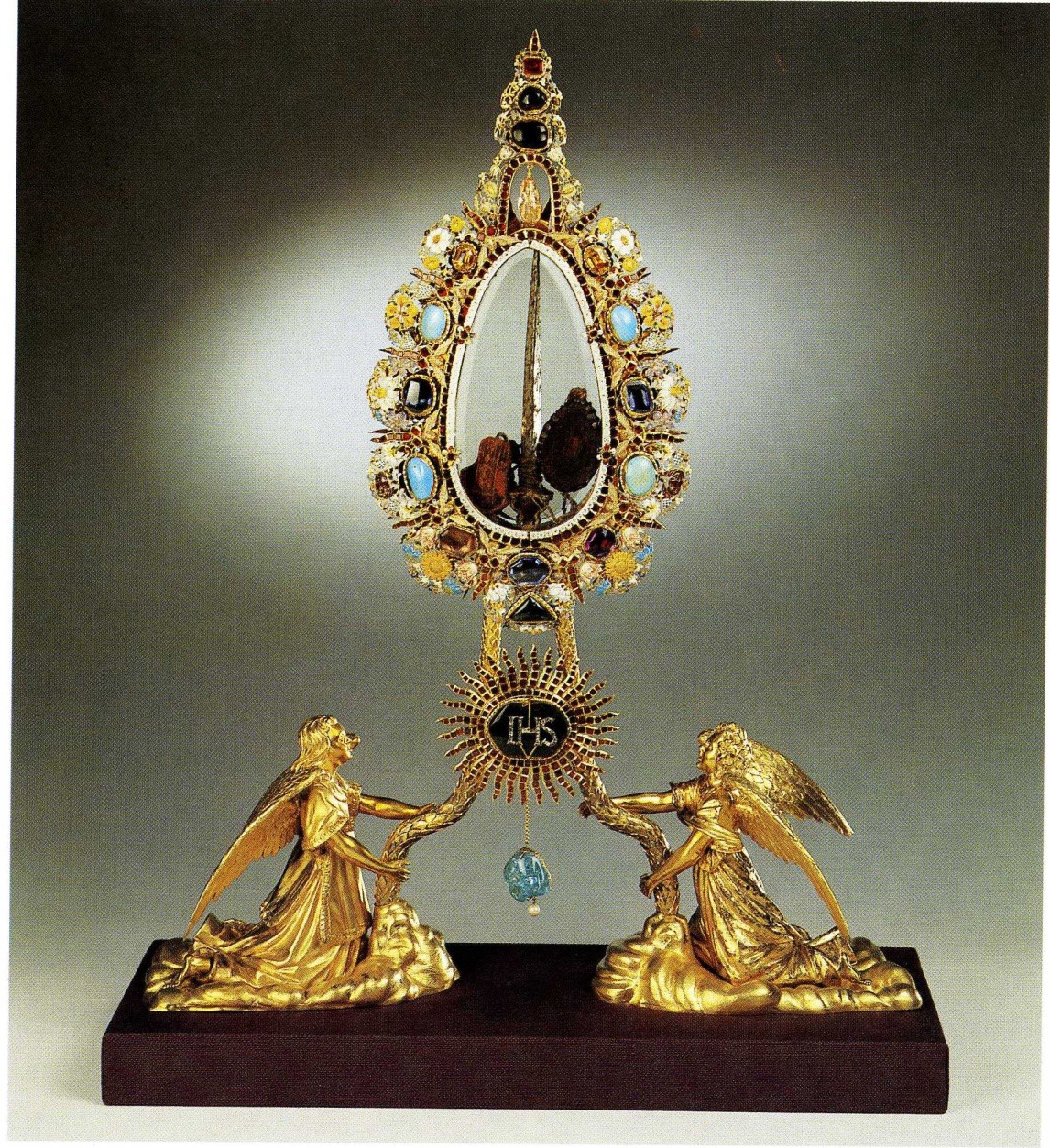 A golden sculpture of two angels holding a jeweled container that displays a nail.