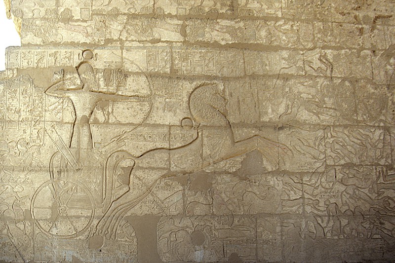 A scene of the battle of Kadesh is carved into a wall made of stone blocks. The king stands in a chariot using his bow and arrow as his horse rears, trampling the tiny people below its hooves.