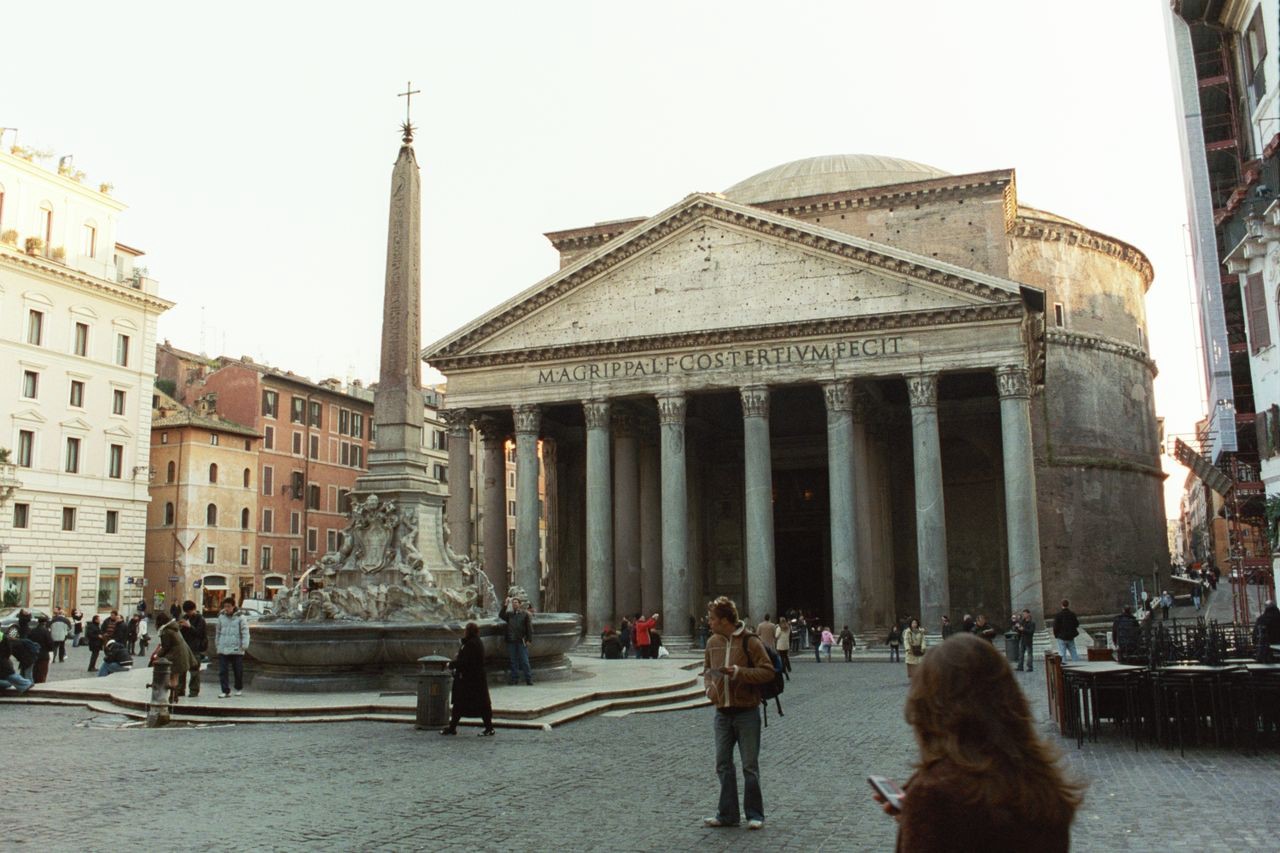 A porch with Corinthian columns holding up a pediment. Behind the pediment is a drum shaped building with a domed roof. In front of the building is a Renaissance fountain built around an obelisk with a cross on top.