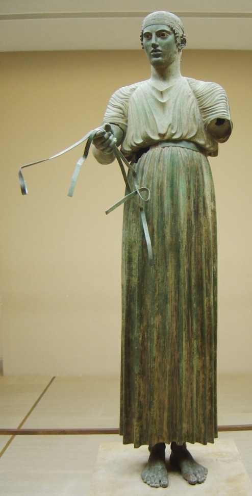 Full figure of the Classical bronze Charioteer. He wears loose clothing tied at the waist and falling to his ankles. His left arm is missing but he holds reins in his right hand.