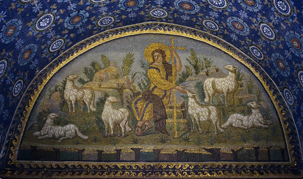 A shepherd with gold and purple clothing sits on a rock surrounded by sheep. He has a gold halo and holds a gold staff shaped like a cross at the top. He wears sandals.