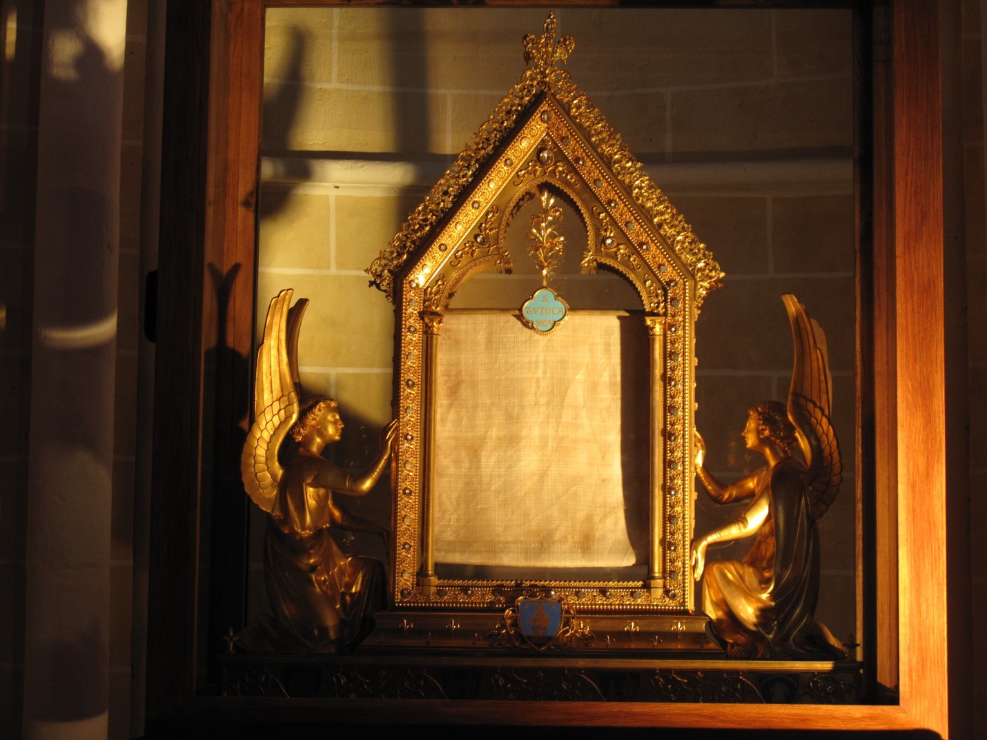 A modern gold container that displays a piece of off-white cloth. The container resembles an altar screen held up by two angels.
