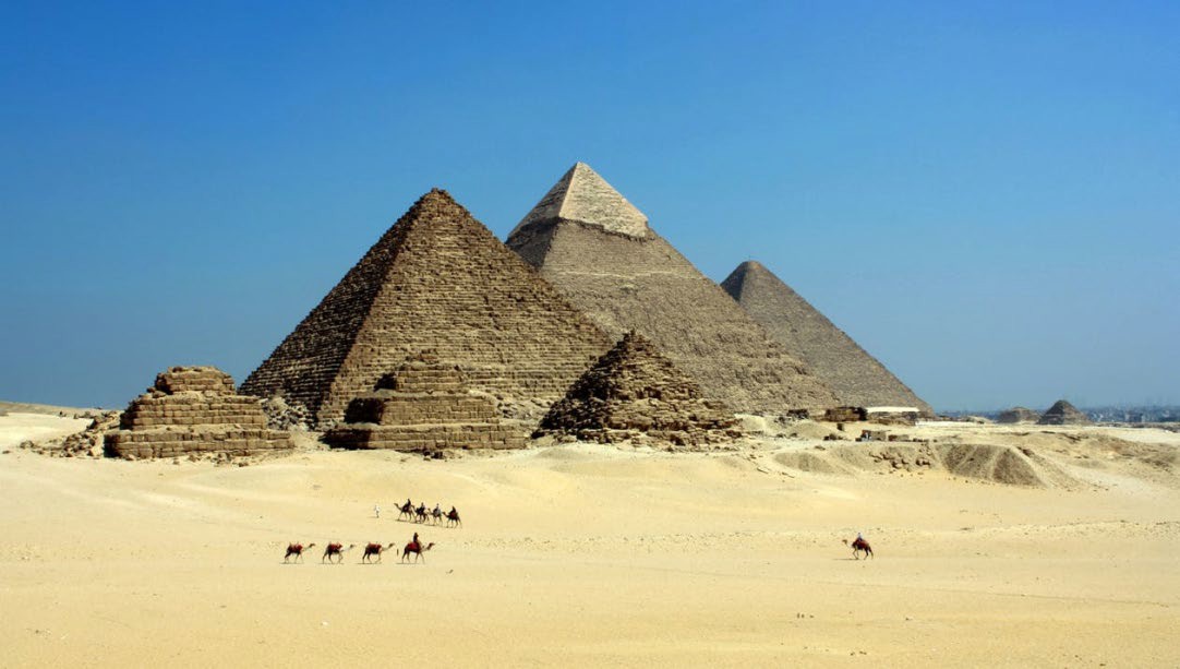 Image of three major pyramids behind three smaller step pyramids. Cales transport people in front of them.