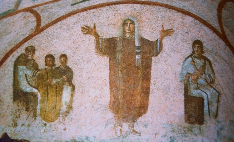 A ceiling fresco with a four spoked wheel. At the end of each spoke is an image from a story. Between the spokes are saints with arms raised in prayer. In the center of the wheel is a shepherd with a sheep over his shoulders. A shelf with skulls is below the painting.