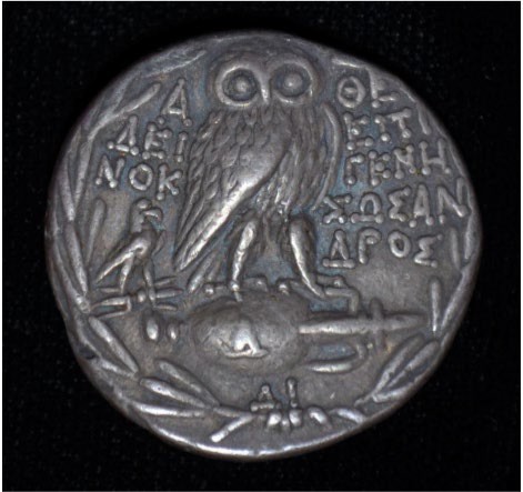 A coin with Athena’s owl as the decoration. There is Greek writing on the sides of the owl.