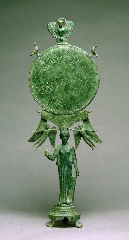 A highly decorative metal mirror held on the head of a figure of Aphrodite. Small, winged figures crown her. The round mirror is topped by another winged figure that resembles an Egyptian Ba image.
