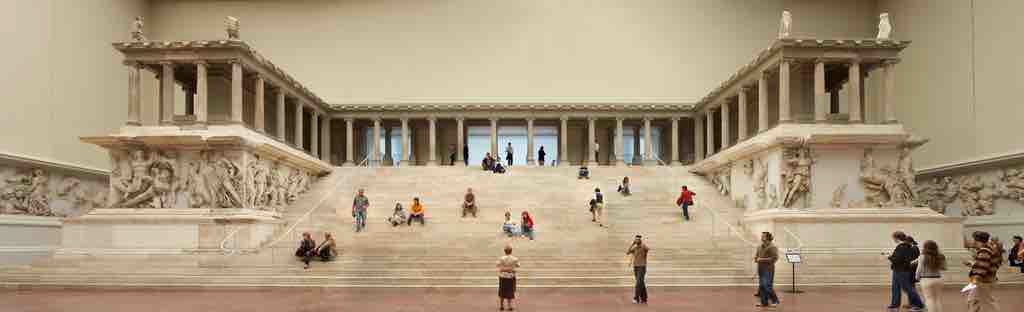 The Alter of Zeus housed inside a museum room. The building is raised on a pedestal and has steps rising to it. The pedestal is covered in a giant frieze that surrounds the building.
