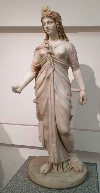 A naturalistic sculpture of a woman with long robes and long hair. She has an emblem of the sun on her forehead.