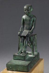 A stylized bronze of a man with a large head. He is seated with a tablet on his lap.