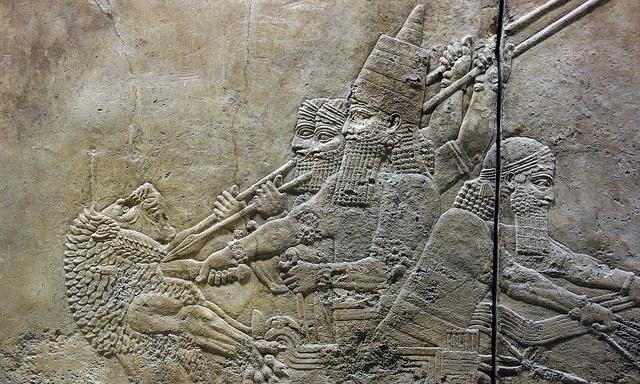 A bas relief of a bearded man wearing a crown plunges a knife into the neck of a male lion while a man drives the chariot he rides and two others hold spears at ready.
