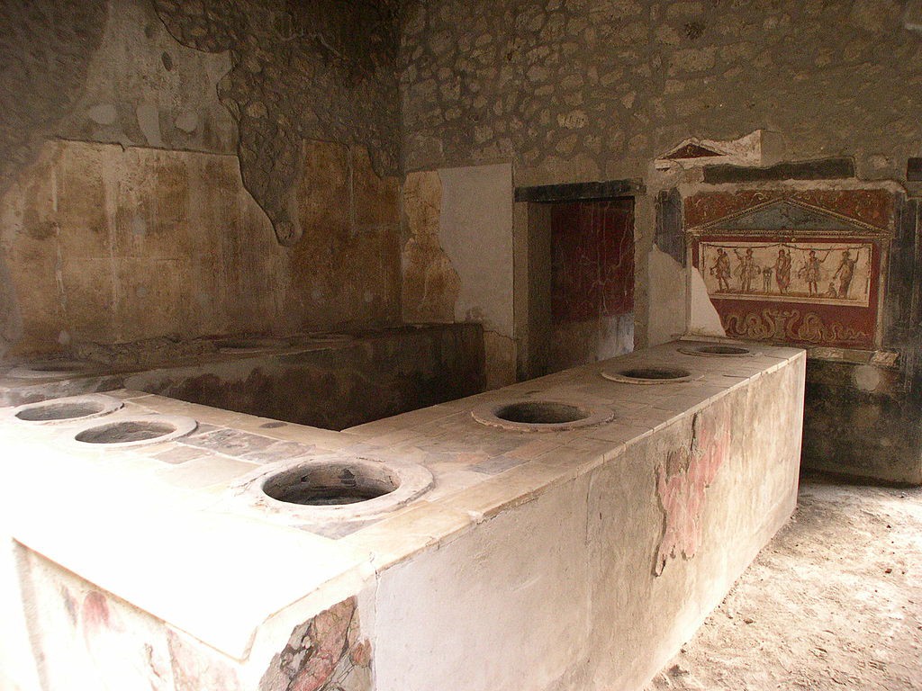 A U-shaped stone counter with round holes indicating places for containers for food. A stone wall with mosaic fragments stands behind it.