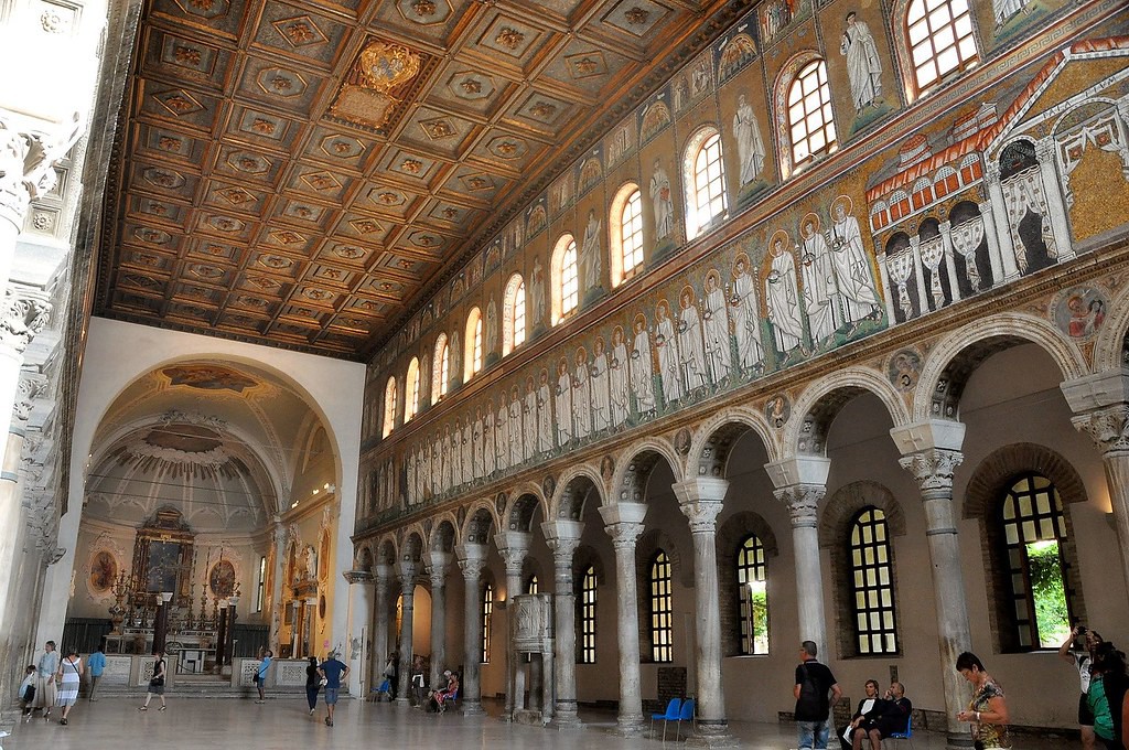 A view of a side aisle with a strip off mosaics above the columns and clerestory windows above that with mosaics of a man beside each.